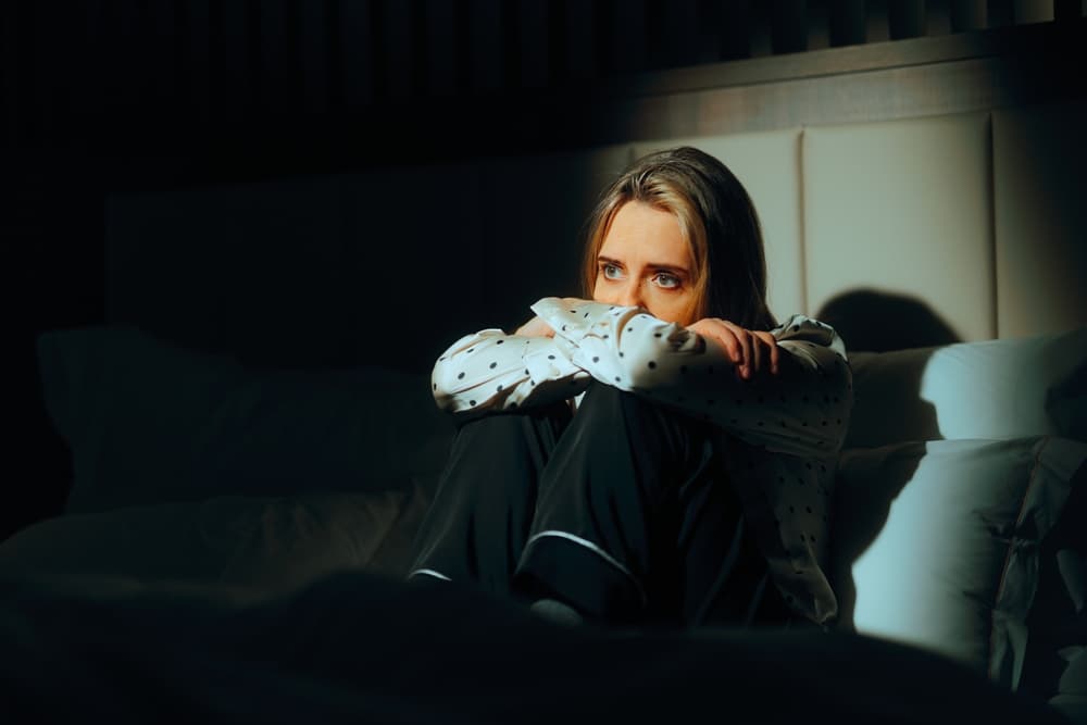 A woman sits alone on a bed in a dark room, knees drawn up, staring into the distance